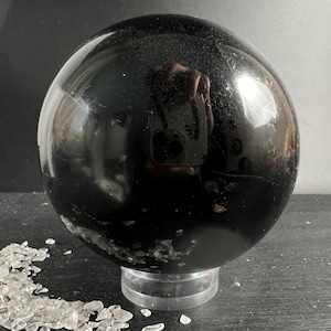 Op de afbeelding: Een grote, gepolijste zwarte bol met een glad, reflecterend oppervlak dat lichtreflecties en een vage weerspiegeling van een camera vertoont. De bol rust op een doorzichtige, ronde kunststof standaard naast kleine, heldere kristalfragmenten.