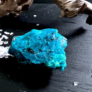 Op de afbeelding: Een levendige, ruwe turkoois edelsteen met donkere insluitsels. De ruw gesneden steen vertoont een rijke, verzadigde blauwgroene kleur. De edelsteen ligt op een donkere houten ondergrond, met kleine witte kristallen en een stuk drijfhout op de achtergrond.