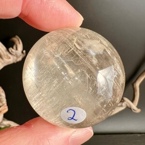 May include: A clear, spherical crystal ball with internal inclusions and a slightly yellow tint. A small, oval sticker with the number "2" is affixed to the bottom. The crystal is held between fingers.