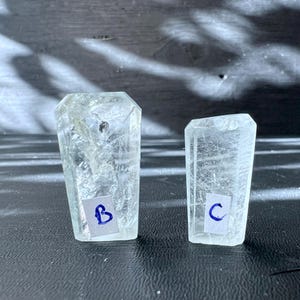 Può includere: Due cristalli di quarzo trasparenti, uno etichettato "B" e l'altro "C", sono esposti su una superficie scura. I cristalli sono entrambi di forma rettangolare e hanno una finitura liscia e lucida.