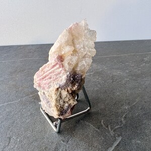 Puede incluir: Un c&uacute;mulo de cristales de lepidolita rosa y blanco sobre un soporte de metal. El cristal tiene una textura rugosa y natural y es de color rosa claro con acentos blancos.
