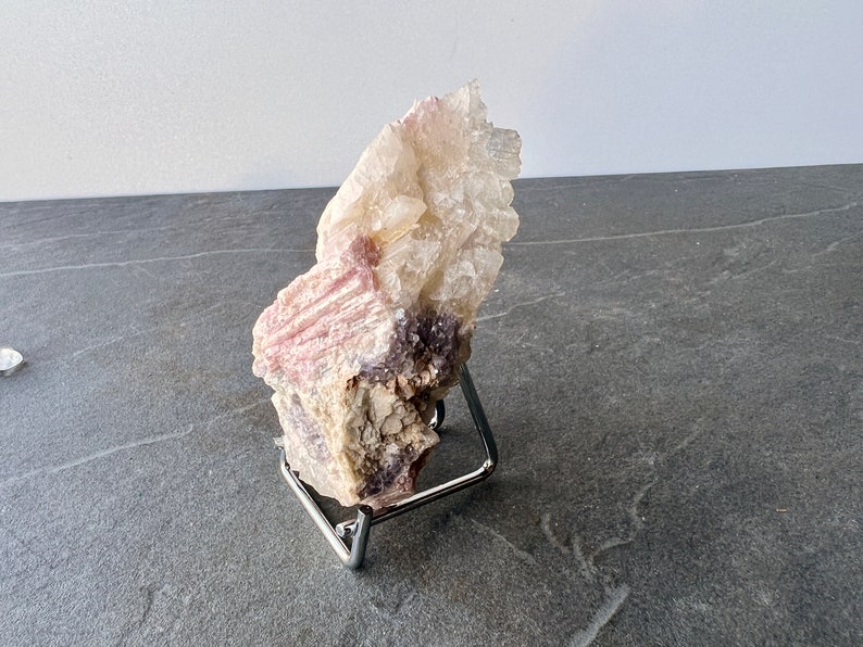 Puede incluir: Un c&uacute;mulo de cristales de lepidolita rosa y blanco sobre un soporte de metal. El cristal tiene una textura rugosa y natural y es de color rosa claro con acentos blancos.