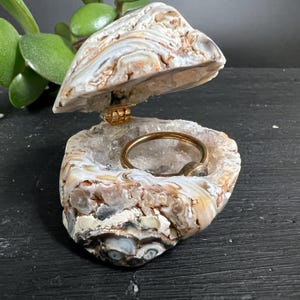 May include: A hinged trinket box crafted from a natural agate geode with a polished, banded exterior in shades of white, tan, and grey. The interior is lined with sparkling clear quartz crystals and contains a simple gold-toned metal ring.