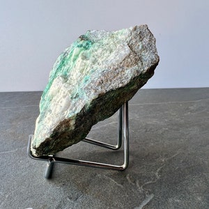 Puede incluir: Una roca grande de forma irregular de color verde y blanco sobre un soporte de metal. La roca tiene una superficie rugosa y texturizada.