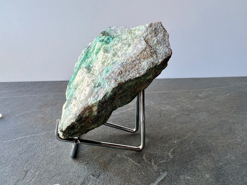 Puede incluir: Una roca grande de forma irregular de color verde y blanco sobre un soporte de metal. La roca tiene una superficie rugosa y texturizada.
