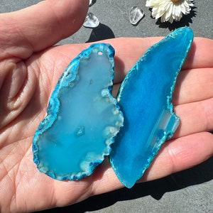 May include: Two blue agate slices with a natural, irregular shape. The agate is translucent and has a light blue colour with white and grey markings.