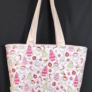 May include: Pink Christmas-themed tote bag with a repeating pattern of Santa Claus, Christmas trees, ornaments, and candy canes. The bag has light pink handles and a matching trim. The background is a checkered pattern.