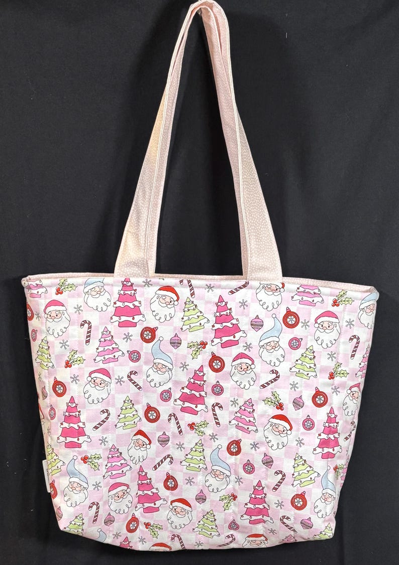 May include: Pink Christmas-themed tote bag with a repeating pattern of Santa Claus, Christmas trees, ornaments, and candy canes. The bag has light pink handles and a matching trim. The background is a checkered pattern.