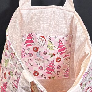 May include: A pink and white Christmas-themed tote bag. The bag features a pocket with Santa Claus, Christmas trees, and candy cane patterns. The interior lining has a white polka dot pattern.