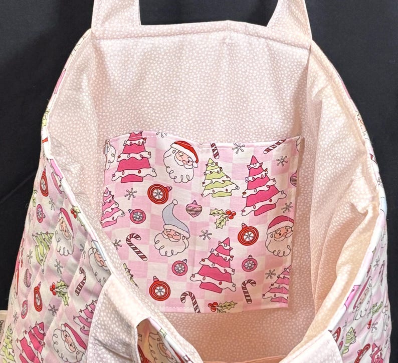 May include: A pink and white Christmas-themed tote bag. The bag features a pocket with Santa Claus, Christmas trees, and candy cane patterns. The interior lining has a white polka dot pattern.