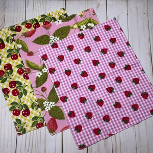 Puede incluir: Tres servilletas de tela estampadas con frutas. Una tiene cerezas sobre un fondo de cuadros amarillos, otra fresas y hojas sobre rosa, y la tercera fresas sobre un estampado de cuadros rosas y blancos.