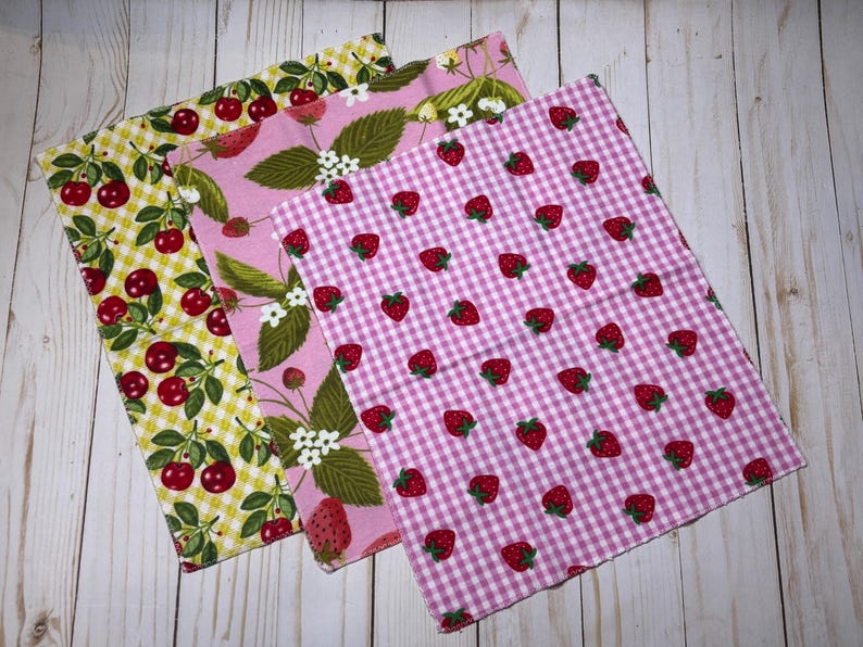 Puede incluir: Tres servilletas de tela estampadas con frutas. Una tiene cerezas sobre un fondo de cuadros amarillos, otra fresas y hojas sobre rosa, y la tercera fresas sobre un estampado de cuadros rosas y blancos.