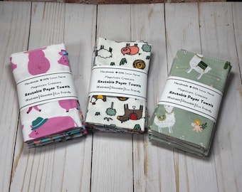 May include: Three stacks of reusable paper towels with different patterns. One stack features pink pigs, another has various farm animals, and the third displays white llamas on a green background. Each stack is wrapped with a label that reads "Reusable Paper Towels."
