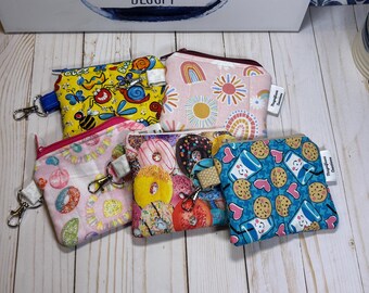 May include: A collection of small, zippered pouches with various colorful patterns. Designs include bees, rainbows, donuts, and cookies with milk. Each pouch has a small strap and a metal clasp.