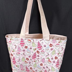 May include: A pink tote bag with a festive Christmas pattern. The fabric features Santa Claus faces, Christmas trees, candy canes, and ornaments on a checkered background. The bag has light pink handles and a matching interior lining.