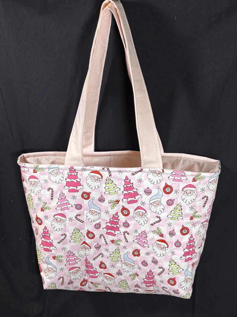 May include: A pink tote bag with a festive Christmas pattern. The fabric features Santa Claus faces, Christmas trees, candy canes, and ornaments on a checkered background. The bag has light pink handles and a matching interior lining.