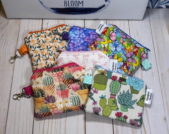 May include: A collection of small, zippered pouches with various fabric patterns. Designs include floral, butterfly, hydrangea, and cactus prints. Each pouch has a coordinating zipper and a small loop with a metal clasp.