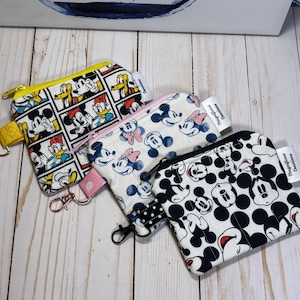 May include: Three small zippered pouches with cartoon character designs. One pouch has a yellow zipper and a Mickey Mouse comic strip pattern. Another has a pink zipper and a Mickey and Minnie Mouse pattern. The third has a black zipper and a Mickey Mouse pattern.