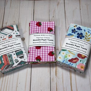 Puede incluir: Tres pilas de toallas de papel reutilizables con diferentes dise&ntilde;os. Una tiene un tema de libros, otra presenta fresas sobre un fondo de cuadros rosas, y la tercera tiene un dise&ntilde;o floral. Cada una est&aacute; etiquetada como "Reusable Paper Towels".