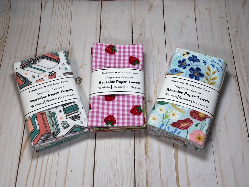Puede incluir: Tres pilas de toallas de papel reutilizables con diferentes dise&ntilde;os. Una tiene un tema de libros, otra presenta fresas sobre un fondo de cuadros rosas, y la tercera tiene un dise&ntilde;o floral. Cada una est&aacute; etiquetada como "Reusable Paper Towels".