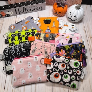 May include: A collection of small, zippered Halloween-themed pouches. Designs include black cats, skeletons, potion bottles, ghosts, and eyeballs. Each pouch has a key ring and is made of fabric with various colors and patterns. A decorative Happy Halloween sign is in the background.