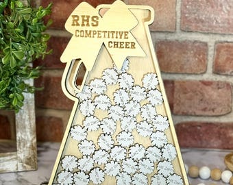 May include: A wooden sign with the text "RHS Competitive Cheer" on it. The sign is shaped like a cheerleader's megaphone and has a small compartment filled with white circles that say "Hit Zero" and "Quiz 1H".