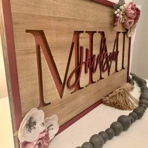 Pu&ograve; includere: Cartello in legno con il nome "MIMI" in grandi lettere bordeaux, con "Hudson" in corsivo sotto. Il cartello &egrave; incorniciato in bordeaux e decorato con fiori finti. Sono presenti anche una ghirlanda di perline grigie e un nappa.