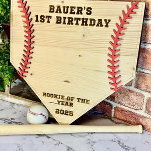 May include: A wooden baseball home plate sign with red stitching and the text "Bauer's 1st Birthday" and "Rookie of the Year 2025". A baseball and a wooden bat are in the foreground.