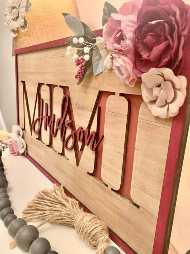Pu&ograve; includere: Cartello in legno con il nome "MIMI" e il nome "Hudson" in corsivo. Il cartello &egrave; decorato con finiture bordeaux, fiori e un nappa di iuta. Lo sfondo &egrave; marrone chiaro.