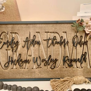 May include: A wooden sign with the word "Grandma" in large, cutout letters. The names "Emily, Aiden, Theodore, Odin, Hudson, Wesley, Reginald" are written in smaller letters below the word "Grandma". The sign has a rustic, farmhouse style and is decorated with a floral arrangement.