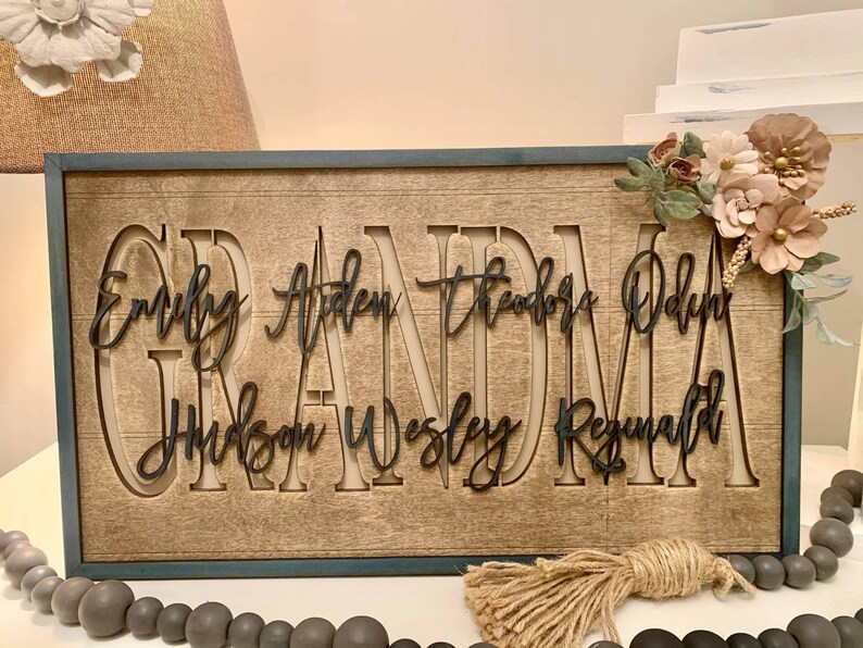 May include: A wooden sign with the word "Grandma" in large, cutout letters. The names "Emily, Aiden, Theodore, Odin, Hudson, Wesley, Reginald" are written in smaller letters below the word "Grandma". The sign has a rustic, farmhouse style and is decorated with a floral arrangement.