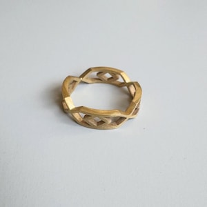 May include: A gold-colored ring with a geometric design. The ring features a series of interconnected diamond and rectangular shapes, creating an openwork pattern. The ring is made of a solid metal and has a matte finish.