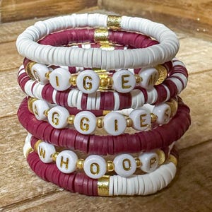 May include: Stack of handmade bracelets in white, burgundy, and gold. The bracelets are made of small, flat, disc-shaped beads. Some bracelets have white beads with gold letter beads spelling out "AGGIE" and "WHOOP".