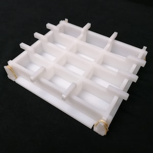 Peut inclure: Moule en plastique blanc avec 9 compartiments rectangulaires. Le moule est ouvert et présente un motif en grille. Le moule est maintenu ensemble par deux élastiques en caoutchouc dorés.