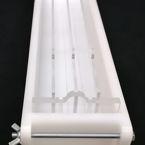 Puede incluir: Molde de pl&aacute;stico blanco con un inserto de acr&iacute;lico transparente. El molde tiene una forma rectangular larga y se mantiene unido por una varilla de metal con tuercas de mariposa. El inserto de acr&iacute;lico tiene cuatro ranuras verticales y un fondo curvo.