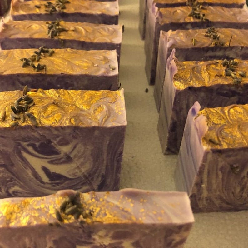 Puede incluir: Primer plano de varias barras de jab&oacute;n hecho a mano. El jab&oacute;n es un remolino de morado y blanco con destellos dorados y botones de lavanda secos en la parte superior.