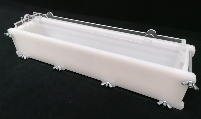 Puede incluir: Un molde de jab&oacute;n rectangular blanco con una tapa de acr&iacute;lico transparente. El molde tiene abrazaderas met&aacute;licas en los lados para asegurar la tapa.