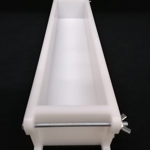 Puede incluir: Molde de jab&oacute;n de pl&aacute;stico blanco con una barra de metal y una tuerca de mariposa. El molde es rectangular y tiene una superficie lisa.