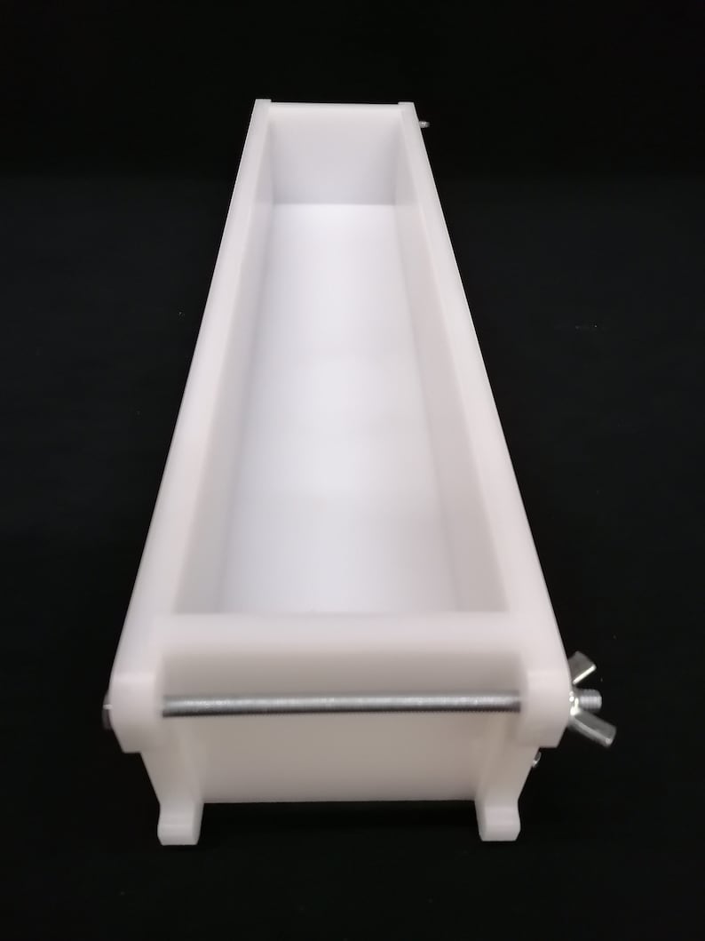 Puede incluir: Molde de jab&oacute;n de pl&aacute;stico blanco con una barra de metal y una tuerca de mariposa. El molde es rectangular y tiene una superficie lisa.