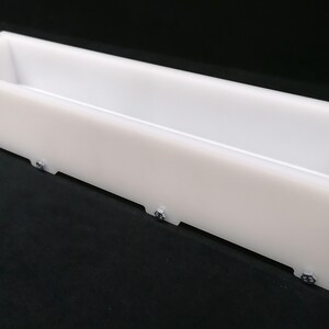 Puede incluir: Un molde de jab&oacute;n rectangular de pl&aacute;stico blanco con un interior liso y un exterior texturizado. El molde se mantiene unido por tornillos.