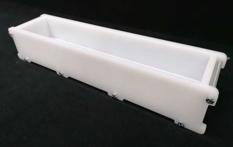 Puede incluir: Un molde de jab&oacute;n rectangular de pl&aacute;stico blanco con un interior liso y un exterior texturizado. El molde se mantiene unido por tornillos.