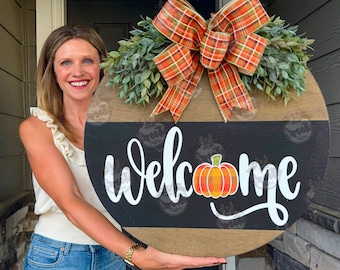 May include: A round wooden door sign featuring a natural wood top and bottom section with a black horizontal centre stripe. The word welcome is written in white script, with a plaid pumpkin replacing the letter o. A large plaid bow sits on top.