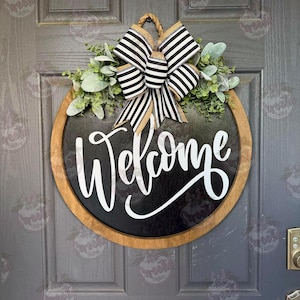 May include: A round wooden door sign with a black center featuring the white script text Welcome. The sign is accented with a large black and white striped bow and green foliage at the top, suspended by a thick twisted rope hanger.