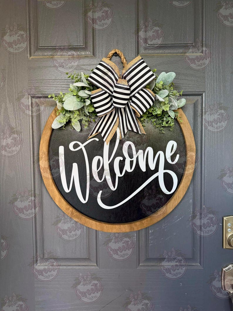 May include: A round wooden door sign with a black center featuring the white script text Welcome. The sign is accented with a large black and white striped bow and green foliage at the top, suspended by a thick twisted rope hanger.