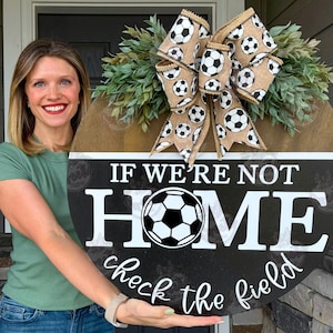 May include: A round wooden sign with the text "IF WE'RE NOT HOME check the field" and a soccer ball graphic. The sign is decorated with a bow featuring soccer ball patterns and greenery.