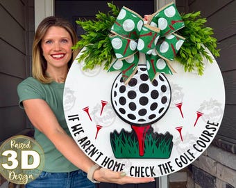 May include: A round white wooden door sign featuring a large 3D golf ball on a red tee, green grass, and a green ribbon with golf ball and tee patterns. The text reads, IF WE AREN'T HOME CHECK THE GOLF COURSE, in black capital letters.