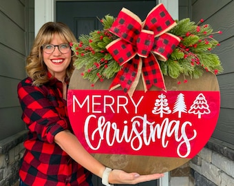May include: A round wooden Christmas sign with a red and white design. The sign features the words "Merry Christmas" in white script, with three white Christmas tree silhouettes. A large red and black buffalo plaid bow and greenery adorn the top.