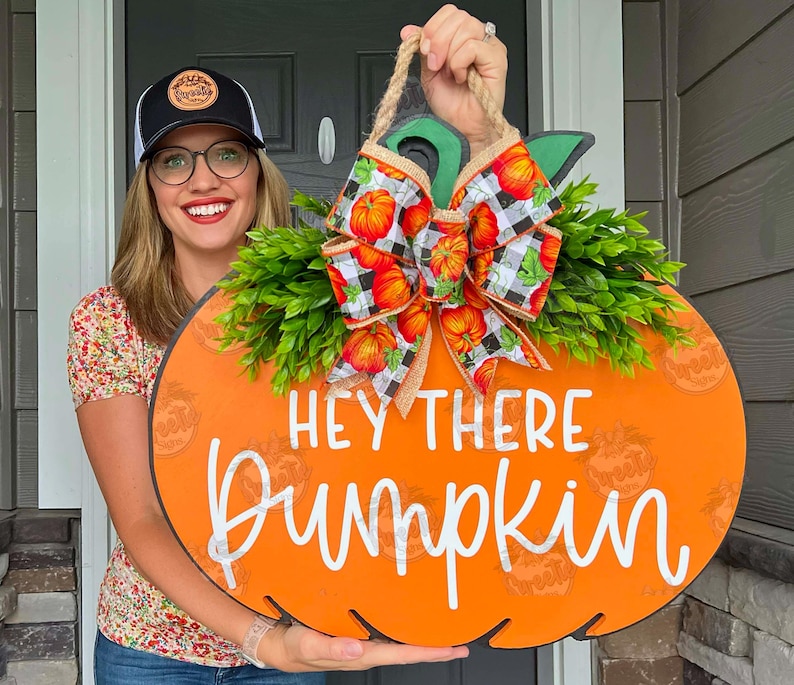 May include: Orange pumpkin shaped wooden door hanger with the text 'Hey there pumpkin' in white. The door hanger is decorated with a green leafy garland and a black and white checkered bow with orange pumpkins on it.