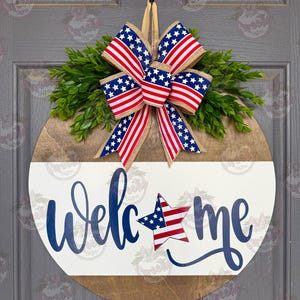May include: A round wooden door sign featuring a natural wood top and a white painted bottom half. The word "welcome" is written in blue script, with a star patterned like the American flag replacing the letter "o". A patriotic bow sits on top.