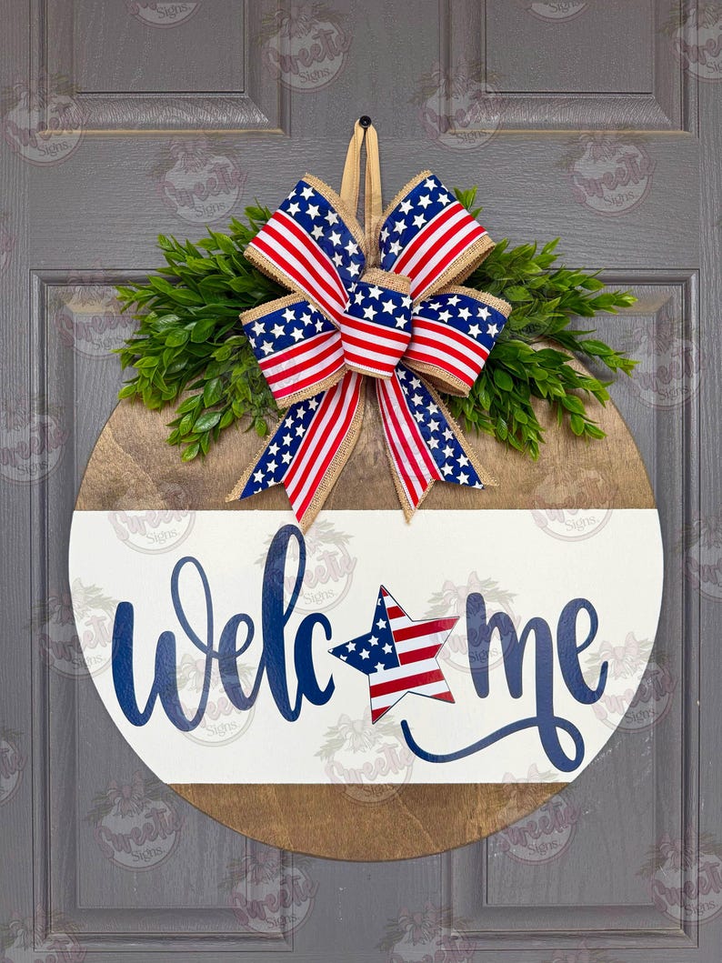May include: A round wooden door sign featuring a natural wood top and a white painted bottom half. The word "welcome" is written in blue script, with a star patterned like the American flag replacing the letter "o". A patriotic bow sits on top.
